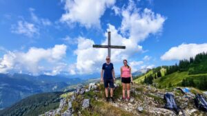 Zwei Vereinsmitglieder stehen neben dem Gipfelkreuz mit toller Aussicht auf die umliegende Landschaft